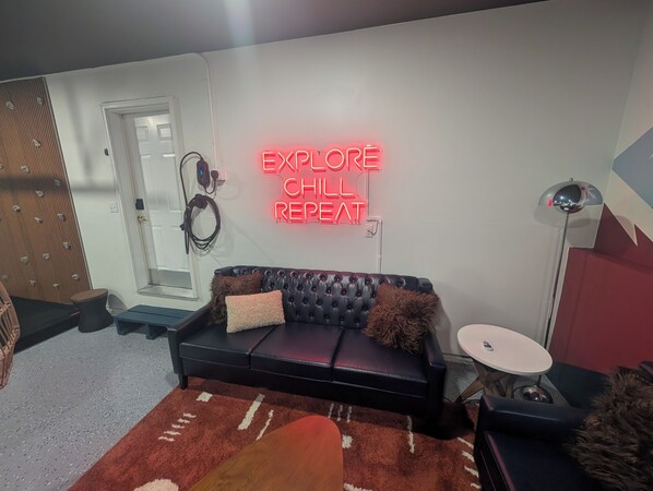 En moderne stue med en sort lædersofa, en neonskilte med teksten 'EXPLORE CHILL REPEAT', et rundt bord og en gulvlampe.