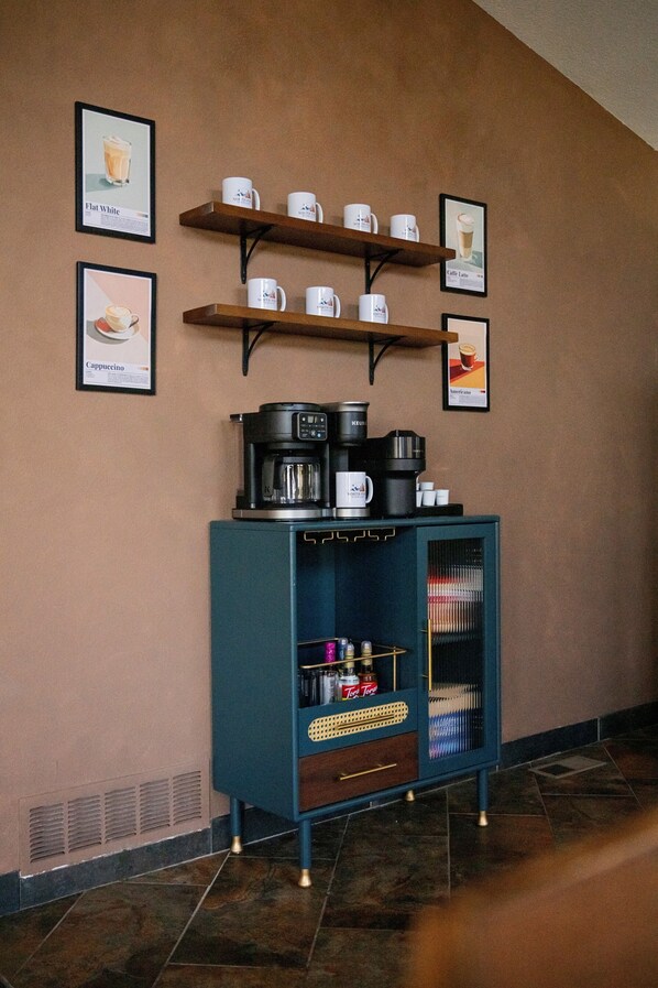Una estación de café con varias cafeteras y tazas en estantes, junto a una pared.