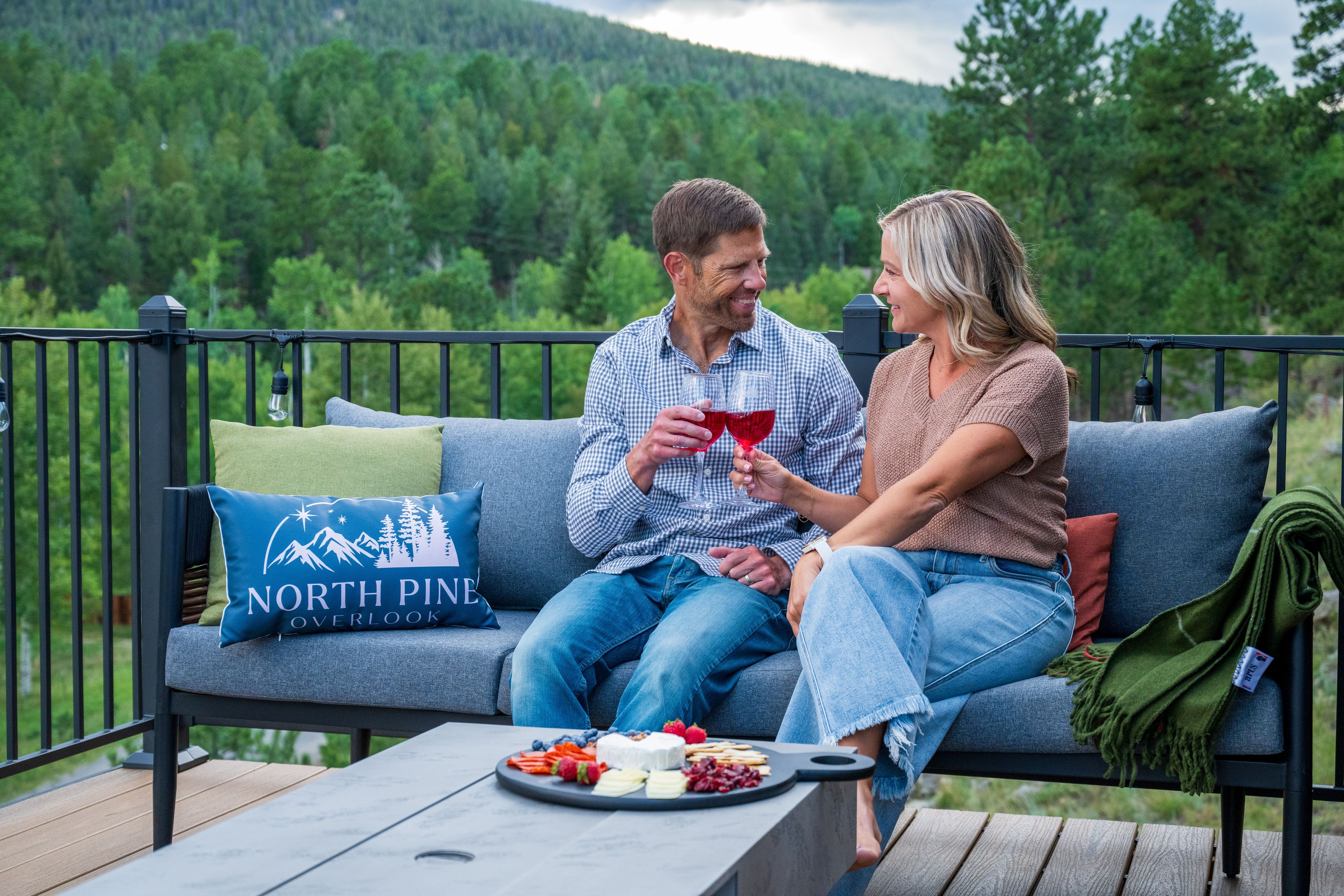 A couple sitting on a grey sofa with a North Pine Overlook pillow, a wooden table with a fruit platter, and a view of a forested area.