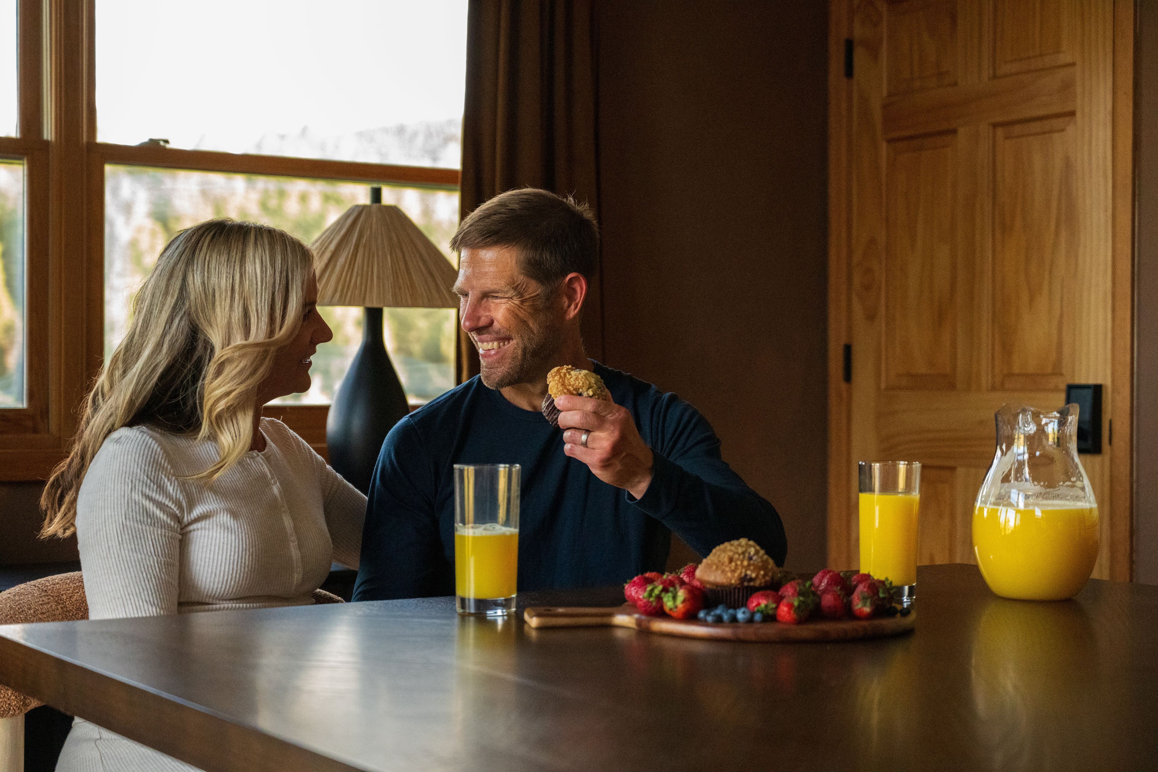 A couple seated at a wooden table with breakfast items, including a muffin and fruit, and two glasses of orange juice.