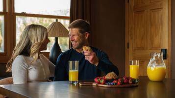 A couple seated at a wooden table with breakfast items, including a muffin and fruit, and two glasses of orange juice.
