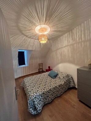 Een kleine, schuine kamer met een bed met patroon, een enkele stoel, een dressoir en een raam.