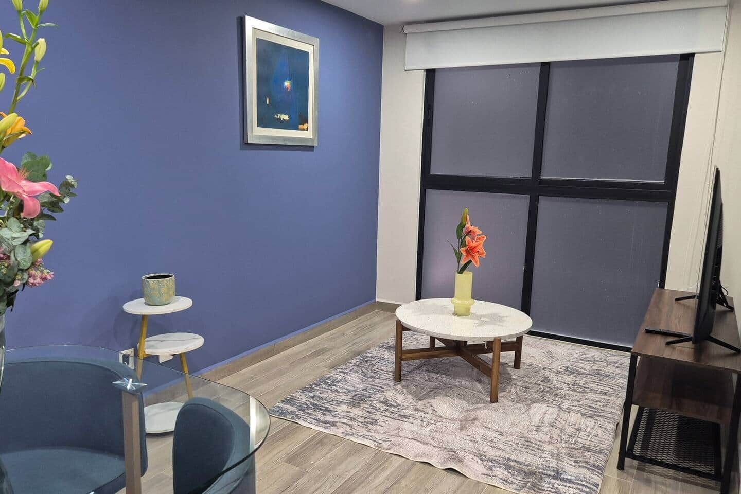 A room with a large window, a round table with a vase of flowers, a small shelf with a cup, and a chair.