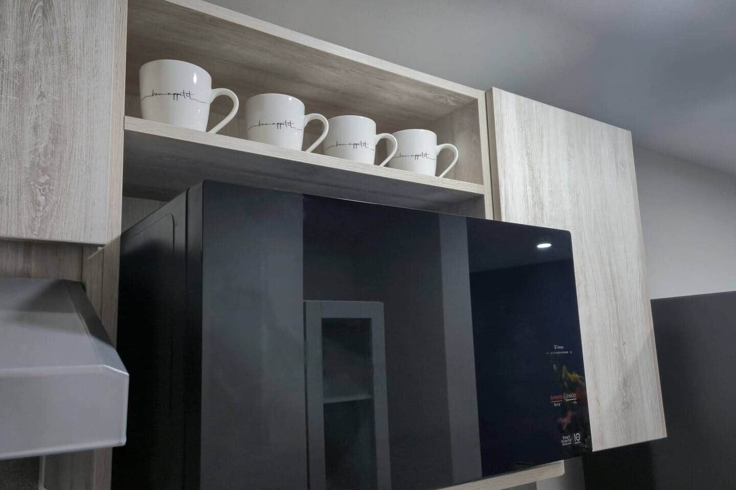 A kitchen with a built-in microwave and a shelf above it holding white mugs.