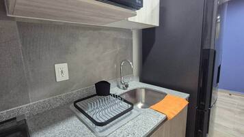 A kitchen with a stainless steel sink, a dish drying rack, a black appliance above the counter, and a refrigerator to the right.
