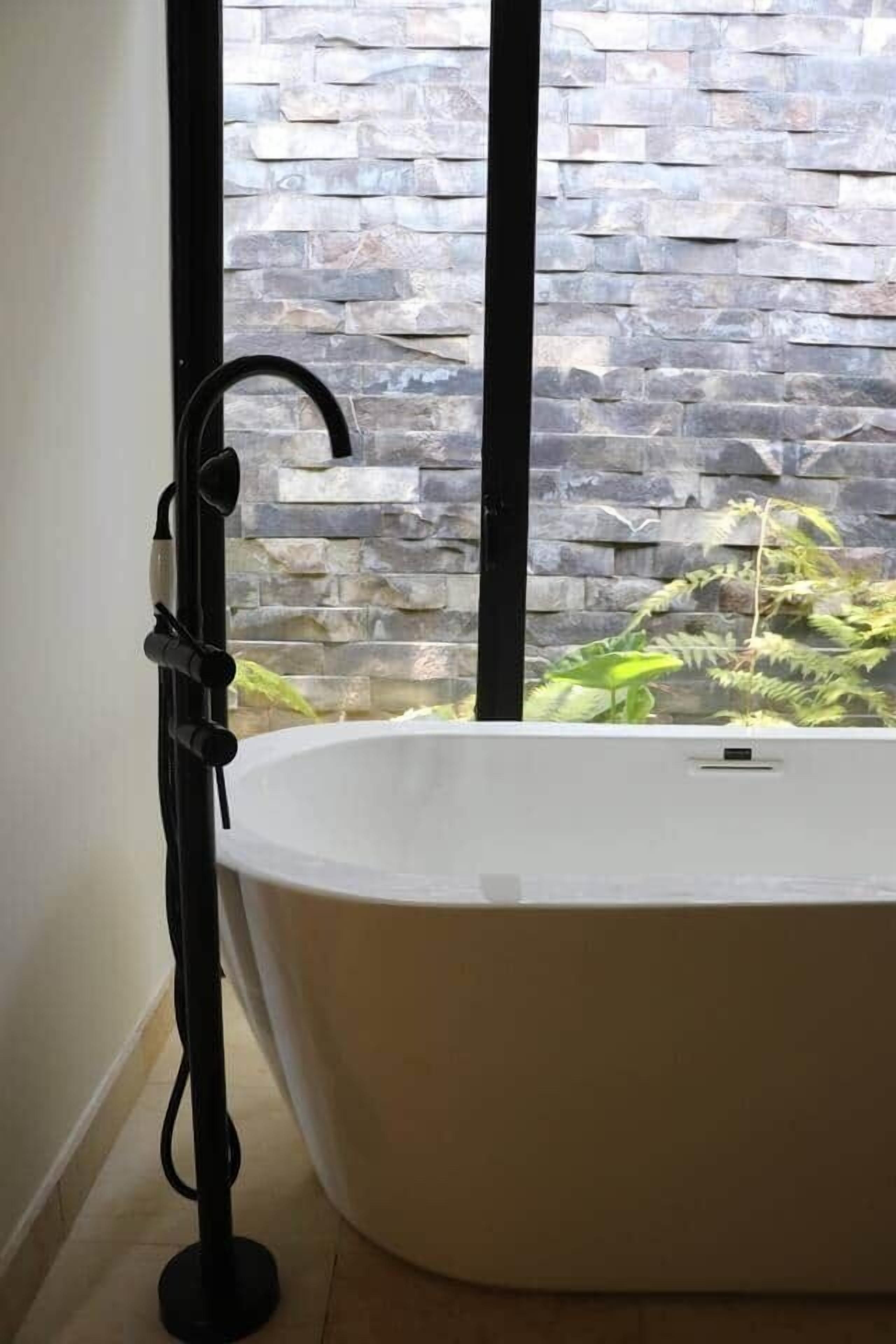 A bathroom with a freestanding bathtub, a black floor lamp, and a stone wall visible through a window.