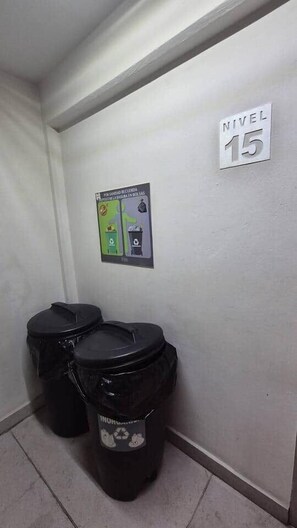 Ein Badezimmer mit zwei schwarzen Mülleimern und einem Recyclingbehälter, einem Schild mit der Aufschrift 'NIVEL 15' und einem Poster an der Wand.