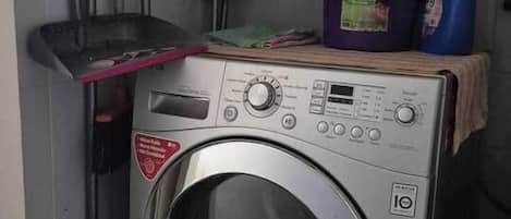 Une machine à laver compacte en acier inoxydable, à chargement frontal, dotée d’un panneau de commande, installée dans une buanderie dont les murs sont recouverts de carreaux et qui comprend une étagère au-dessus, où sont rangés les produits de nettoyage.