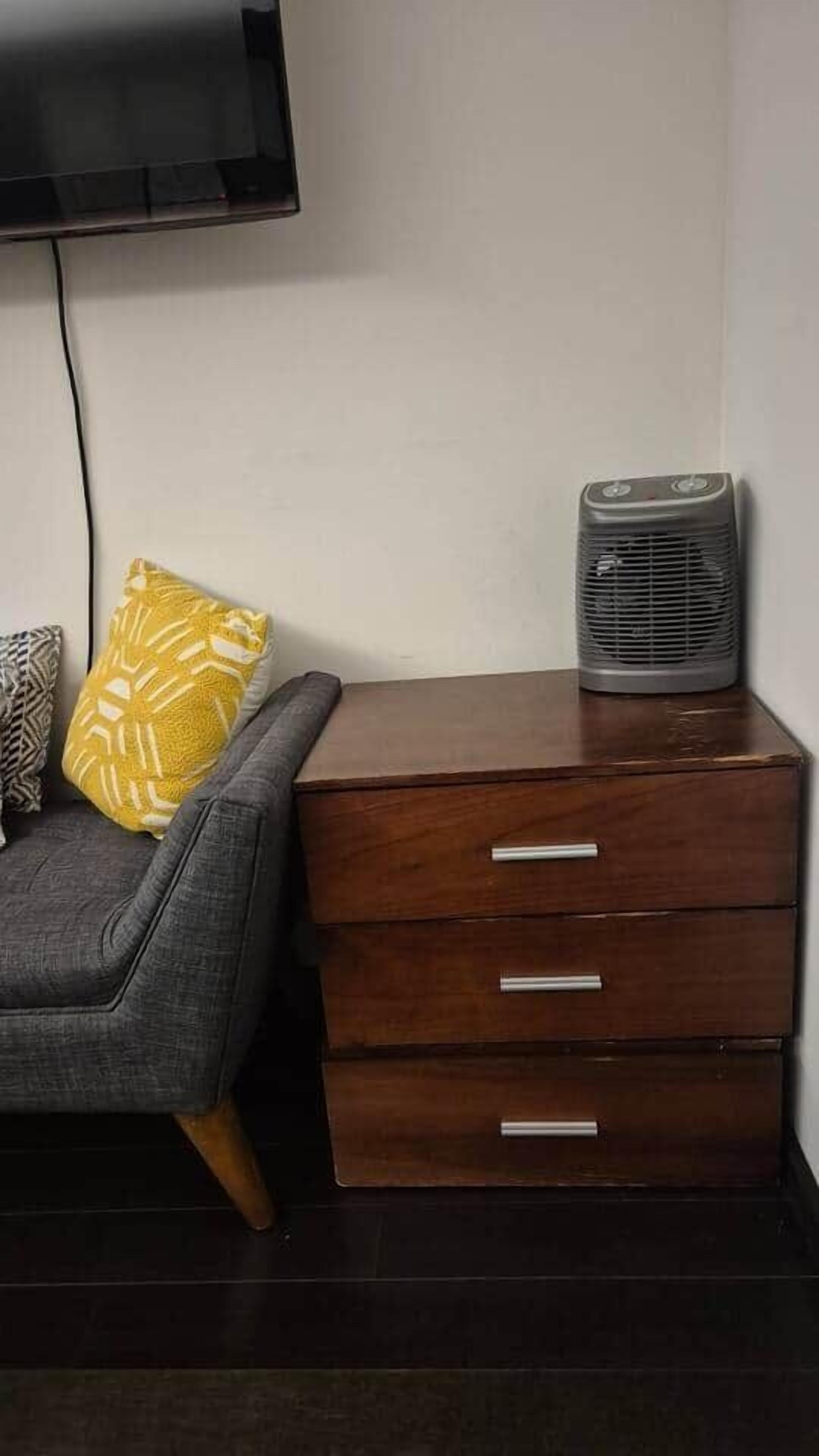 A room with a grey sofa, a wooden chest of drawers, a black heater, and a television mounted on the wall.