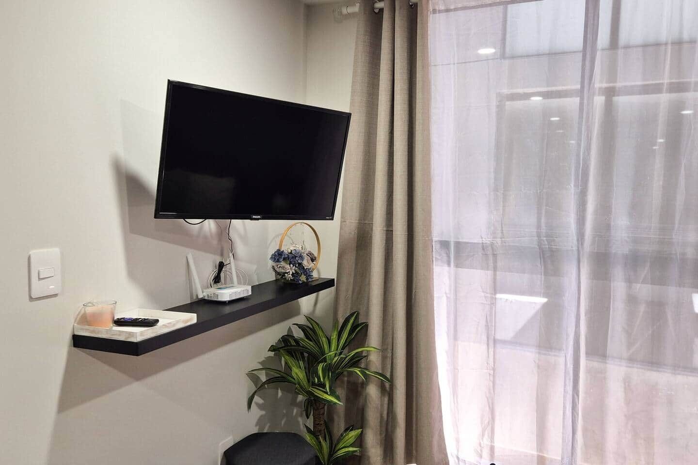 A modern room with a mounted flat-screen TV, a small table with a remote and a projector, a potted plant, and a window with sheer curtains.