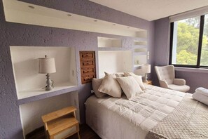 Une chambre à coucher avec un lit, une chaise, une table de chevet et une étagère avec une lampe.