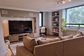 A modern living room with a large flat-screen TV, a sofa, a desk, and a bookshelf. - Fabian (Rio de Janeiro) (Mexico City)