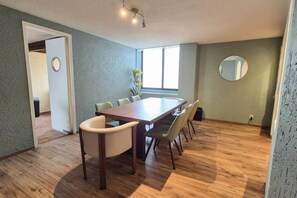 A dining area with a wooden table and chairs, a potted plant, a round mirror, and a window providing natural light. - Fabian (Rio de Janeiro) (Mexico City)