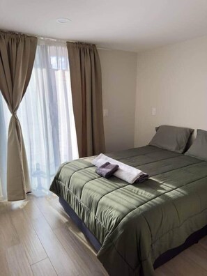 A single bed with a green bedspread, a pair of folded towels, and a window with beige curtains. - Erick Insurgentes A 606 (Mexico City)