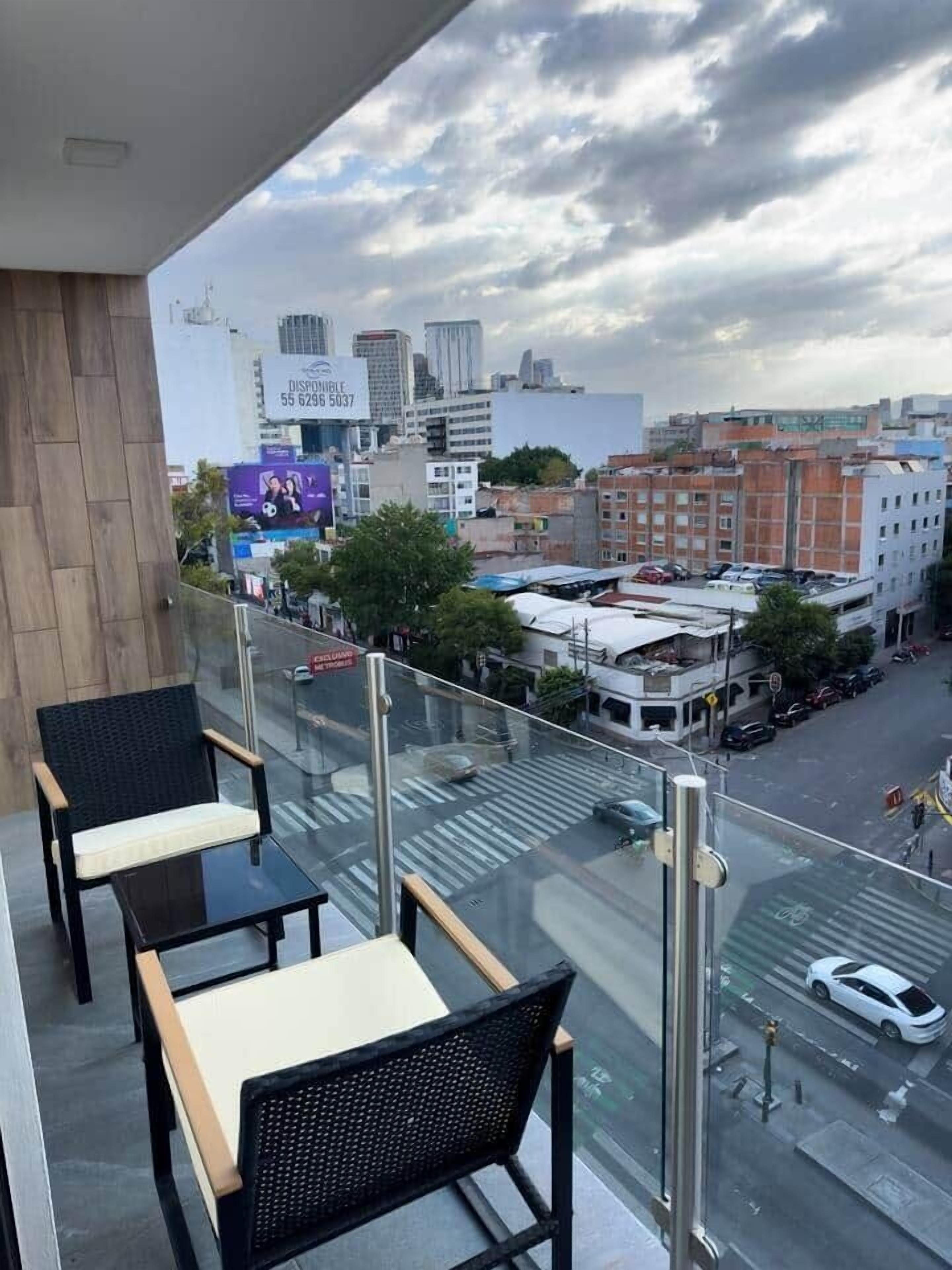 A balcony with two chairs and a table, overlooking a city street with buildings and a billboard.
