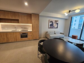 A modern kitchen with wooden cabinets, a built-in oven, and a marble countertop. There is a dining area with a round table and chairs, and a sofa. A large window provides a view of the city. - N Miami Ave - Suites (Miami)