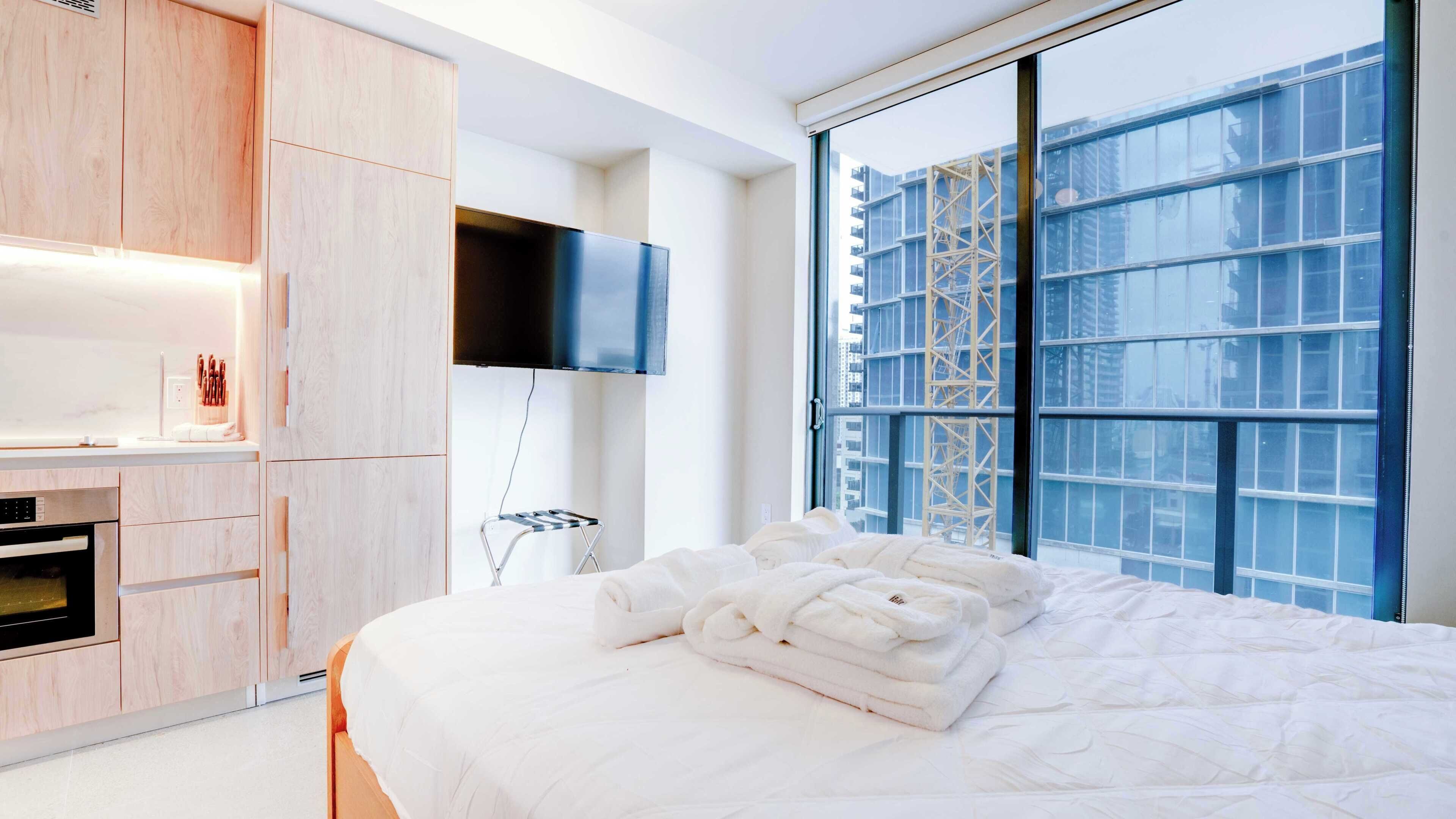 Una habitación de hotel moderna con una cama grande, un televisor de pantalla plana, horno integrado y vistas a la ciudad a través de amplios ventanales.