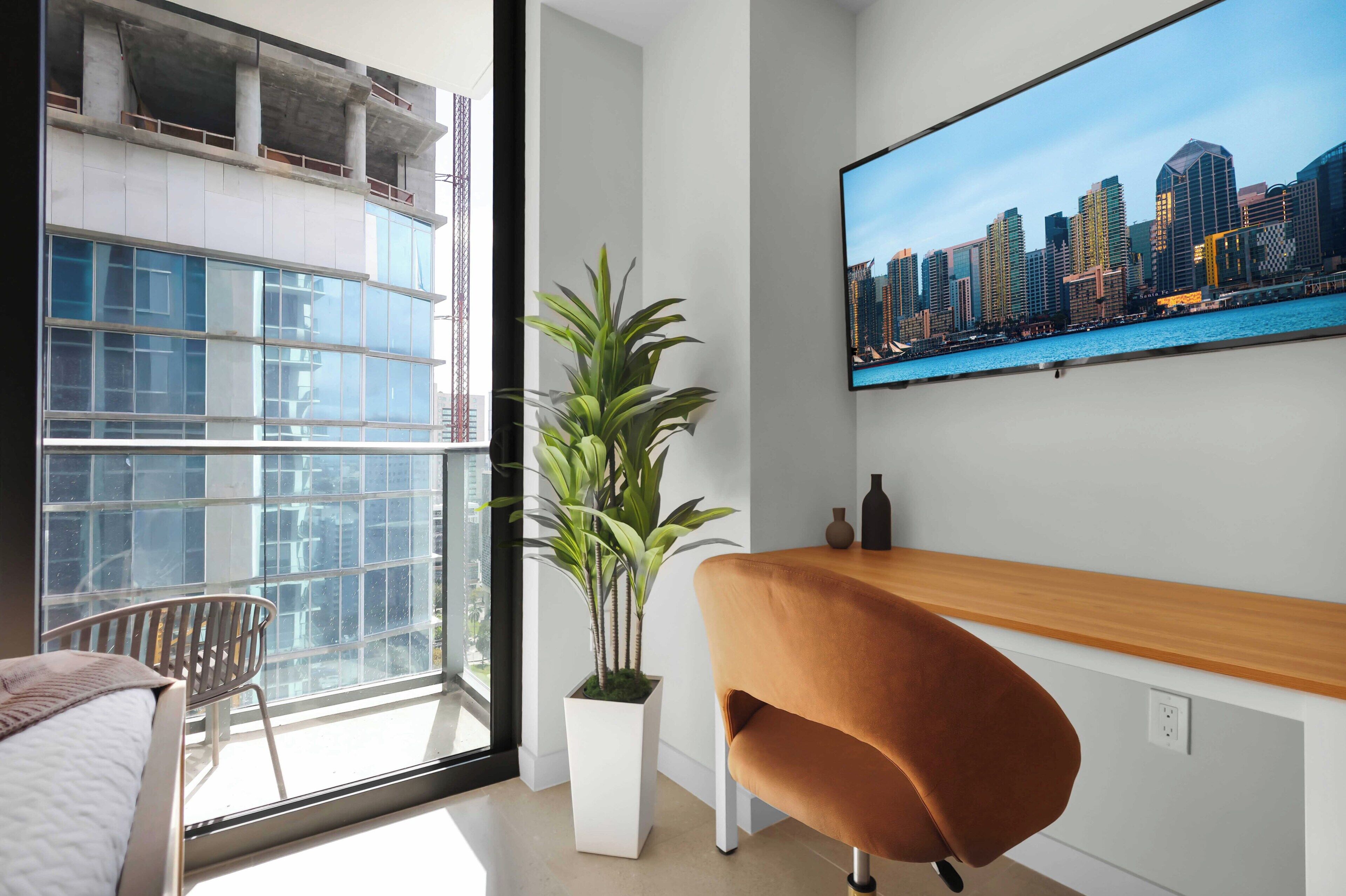 A modern room with a large window, a potted plant, a wooden desk, a chair, and a cityscape painting.