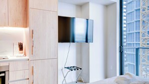 Una habitación de hotel moderna con un televisor de pantalla plana en la pared, un taburete plegable, una mesita de noche con lámpara y una ventana con vista a un edificio.