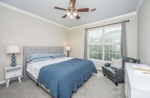 A bedroom with a bed, bedside tables, a desk, a chair, a ceiling fan, and a window with curtains.