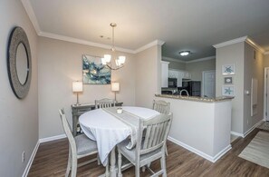 A dining area with a round table, four chairs, a chandelier, and a kitchen in the background.