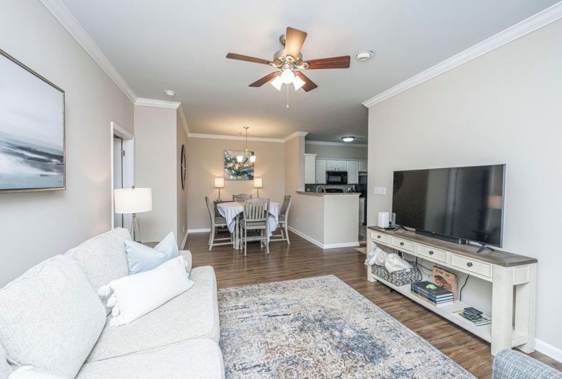 A living room with a sofa, a TV, a ceiling fan, a dining area, and a kitchen in the background.