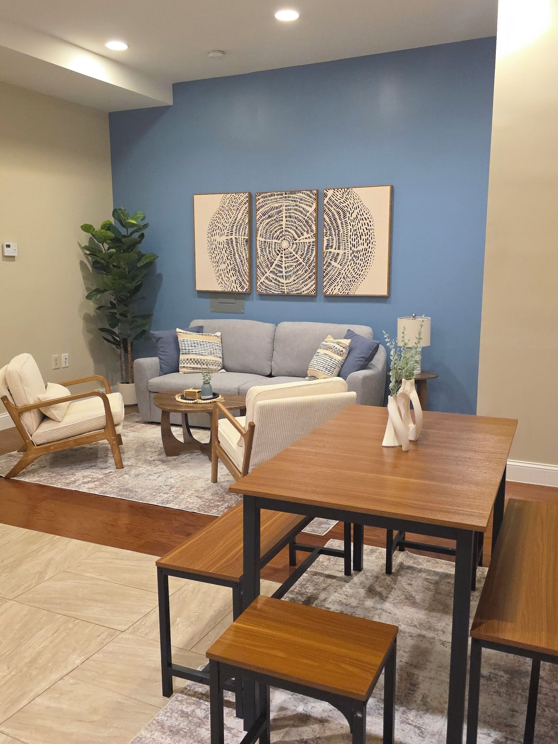 A living room with a sofa, two armchairs, a coffee table, and a dining table. There are three wall art pieces above the sofa.