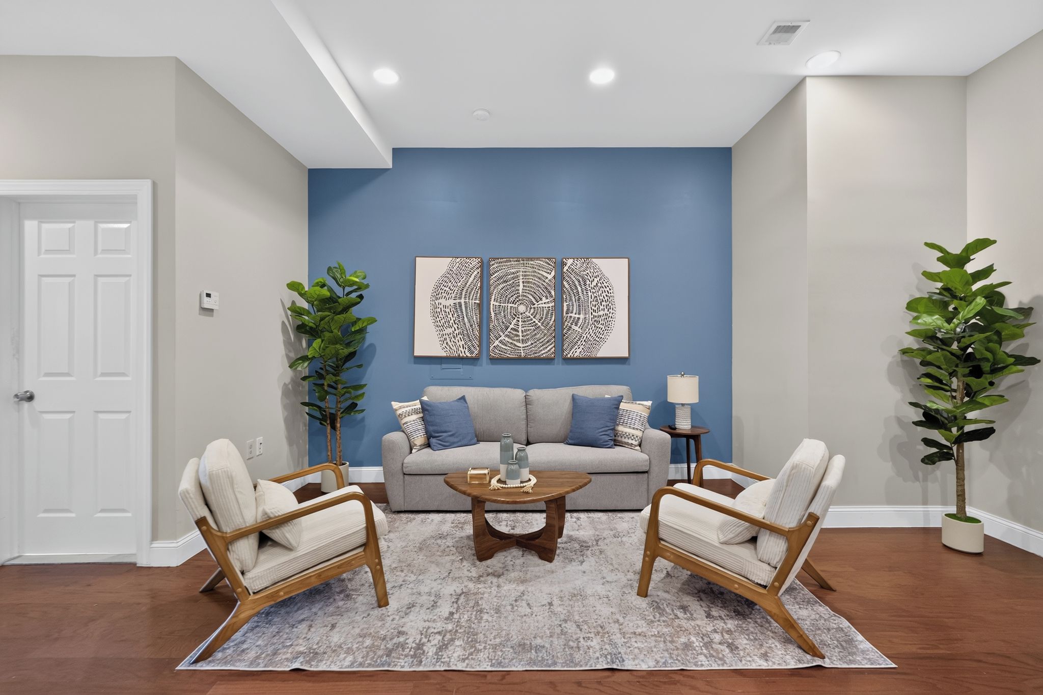 A living room with a blue accent wall, a grey sofa, two armchairs, a coffee table, and two potted plants.