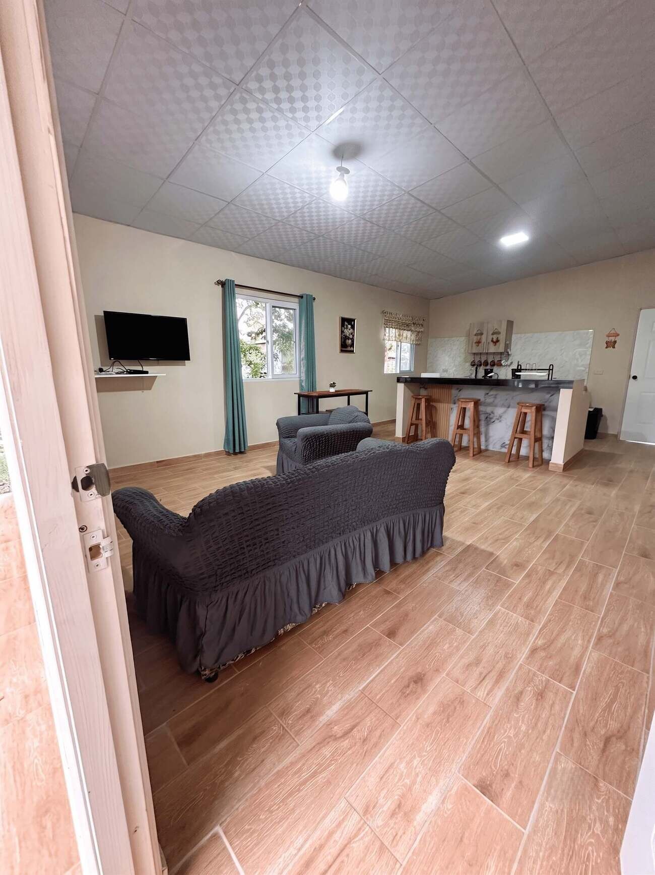 A spacious living area with a sofa set, a flat-screen TV mounted on the wall, and a kitchen area with bar stools.