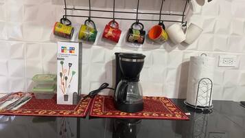 Una cuina amb un taulell, una cafetera, un suport penjant per a tasses i un conjunt d'estris.