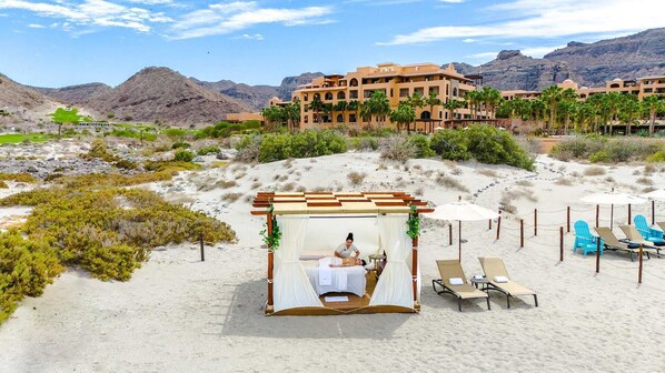 A desert resort with a massage tent, lounge chairs, and a building with palm trees in the background.