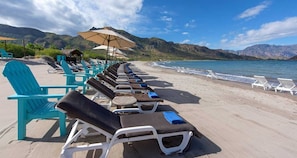 A beach with rows of sun loungers and umbrellas, a clear sky, and mountains in the background.