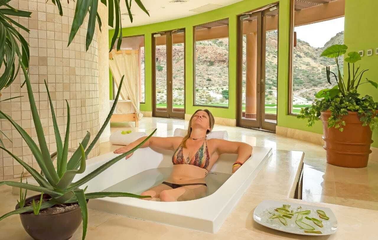 Une femme se détend dans une baignoire, avec vue sur un paysage montagneux grâce à de grandes fenêtres.