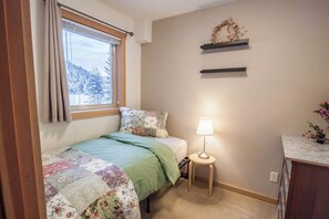 A bedroom with a bed, a bedside table with a lamp, a window with curtains, and a wall-mounted shelf with decorative items. - From cozy single rooms to spacious suites, we have something for everyone. (Seward)