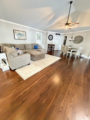 A living room with a sectional sofa, hardwood flooring, a dining area with chairs, a ceiling fan, and a wall clock. - Cozy Lake Escape on Weiss Lake (Cedar Bluff)
