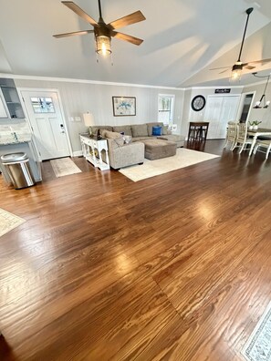 A spacious living room with a large sectional sofa, wooden flooring, ceiling fans, and a dining area with chairs. - Cozy Lake Escape on Weiss Lake (Cedar Bluff)
