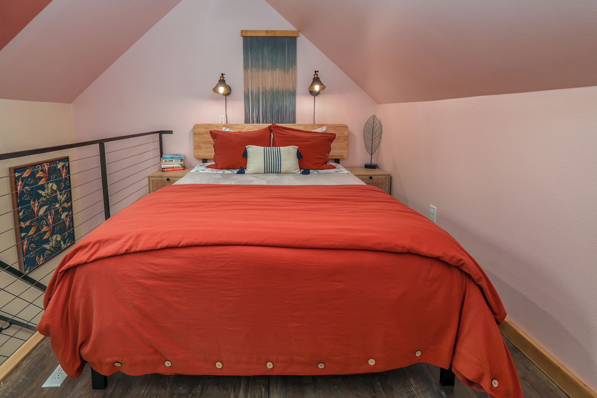 Une chambre avec un plafond en pente, un lit recouvert d’une couverture rouge, deux lampes de chevet, une table de chevet avec des livres et une fenêtre avec des rideaux.