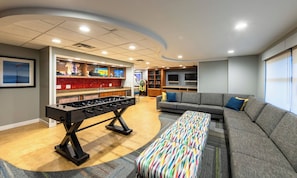 A room with a foosball table, a sofa, a colorful ottoman, and a bar area with a television and shelves.