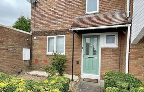 Une maison à deux étages, construite en briques, avec une porte verte, des fenêtres à cadre blanc et un petit jardin parsemé de fleurs jaunes.