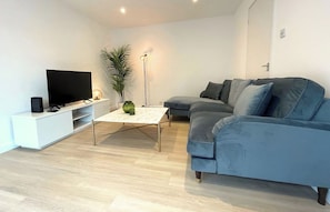 Un salon moderne avec un téléviseur à écran plat, une console blanche, un canapé bleu et une lampe sur pied.