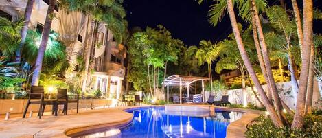 A nighttime swimming pool area with a curved design, surrounded by palm trees and outdoor seating.