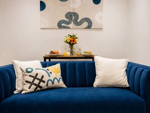 Ein blaues Sofa mit zwei Zierkissen, ein kleiner Tisch mit Blumen in einer Vase und eine Wand mit abstrakter Kunst.