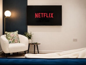 Ein modernes Wohnzimmer mit einem weißen Sessel, einem kleinen Beistelltisch mit einer Pflanze und einem an der Wand montierten Fernseher, der 'NETFLIX' anzeigt.