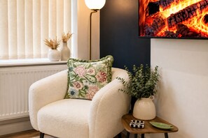 Ein gemütliches Zimmer mit einem weißen Sessel, einem Zierkissen, einem kleinen Tisch mit einer Vase und einem karierten Gegenstand, sowie einem an der Wand hängenden Kunstwerk mit Feuerdarstellung.