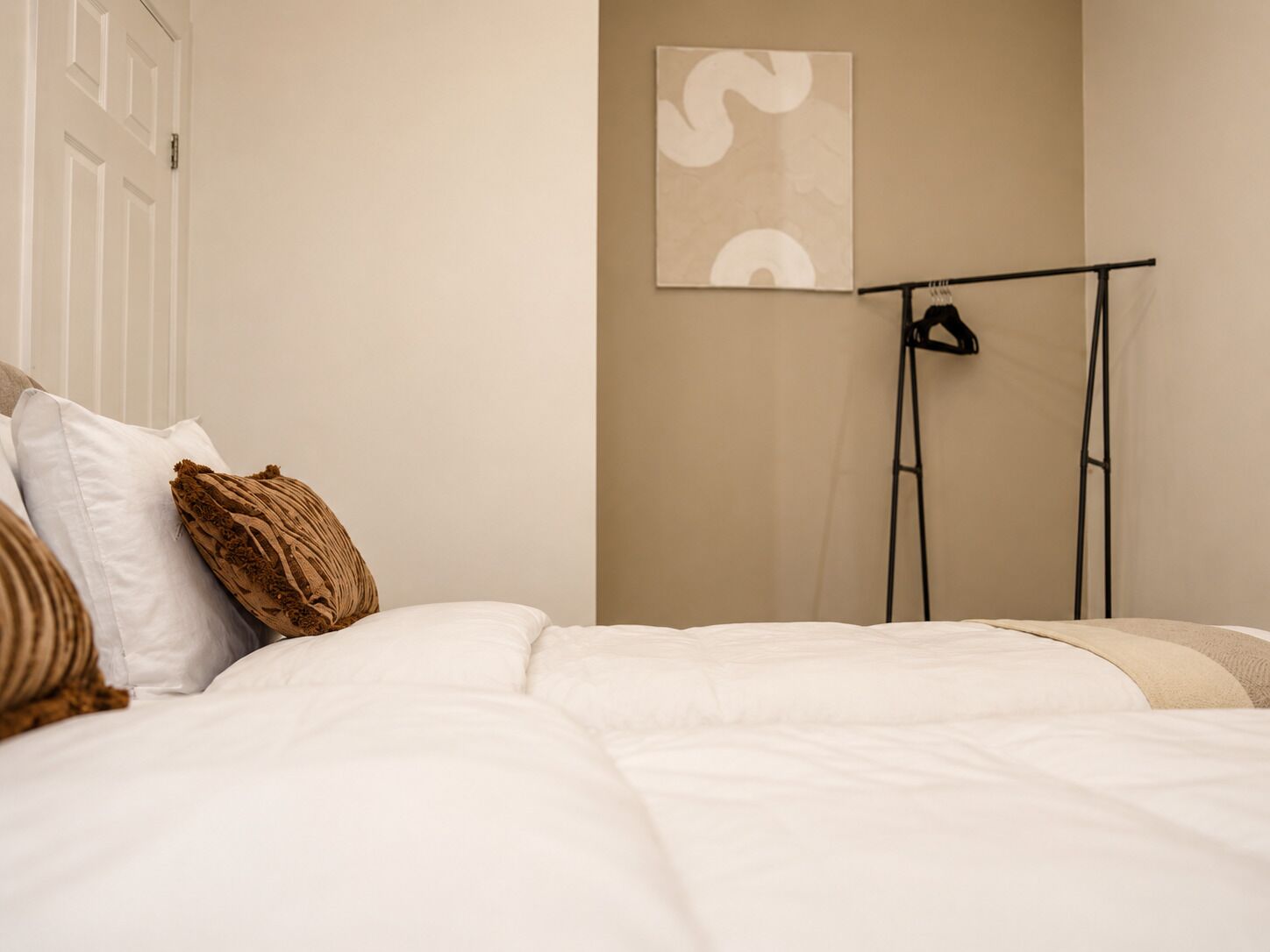 Un dormitorio con cama, almohadas, una obra de arte en la pared y un perchero negro sobre un soporte.