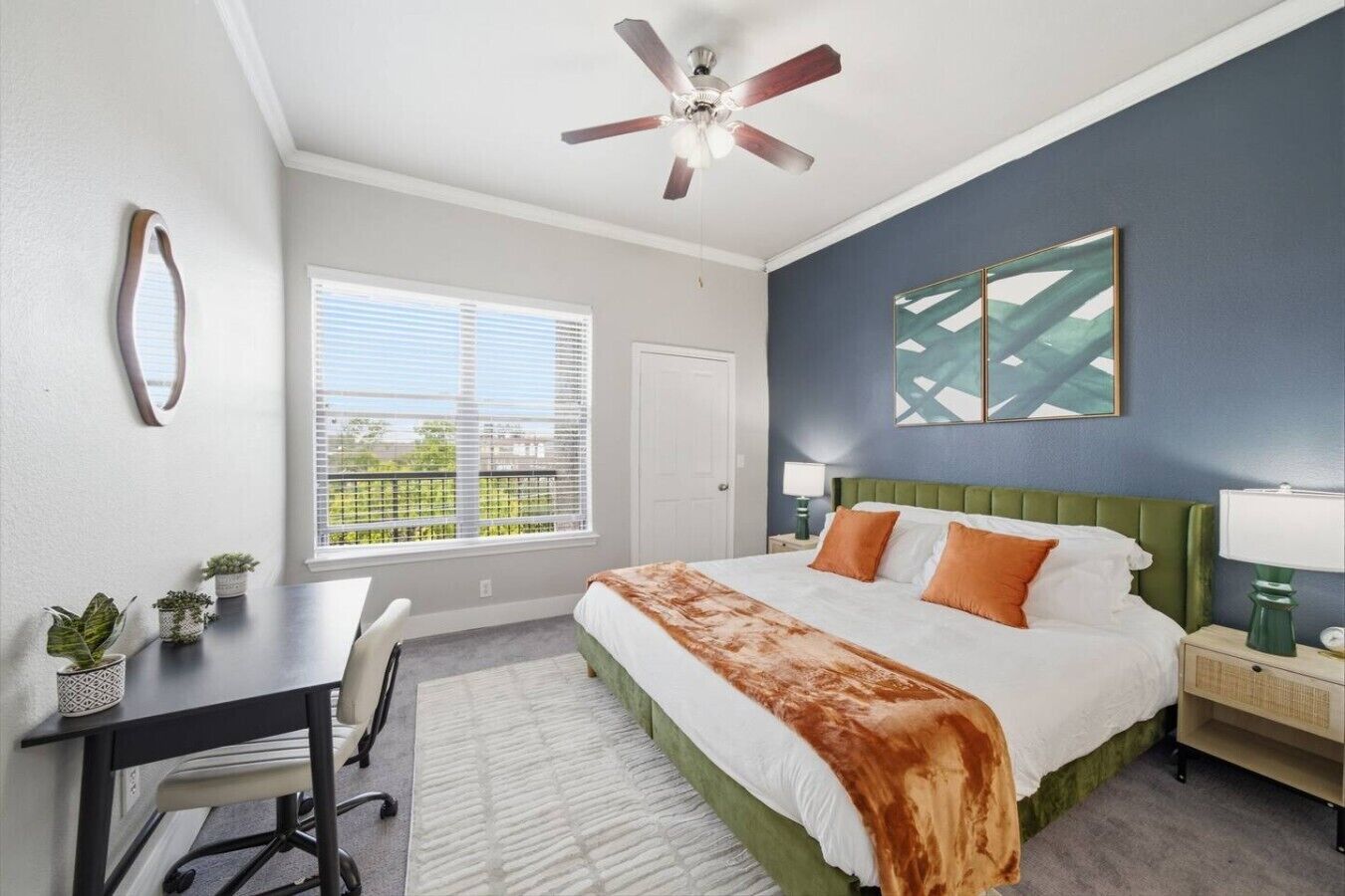 A bedroom with a bed, a desk, a chair, a ceiling fan, a window with blinds, and a wall-mounted artwork.