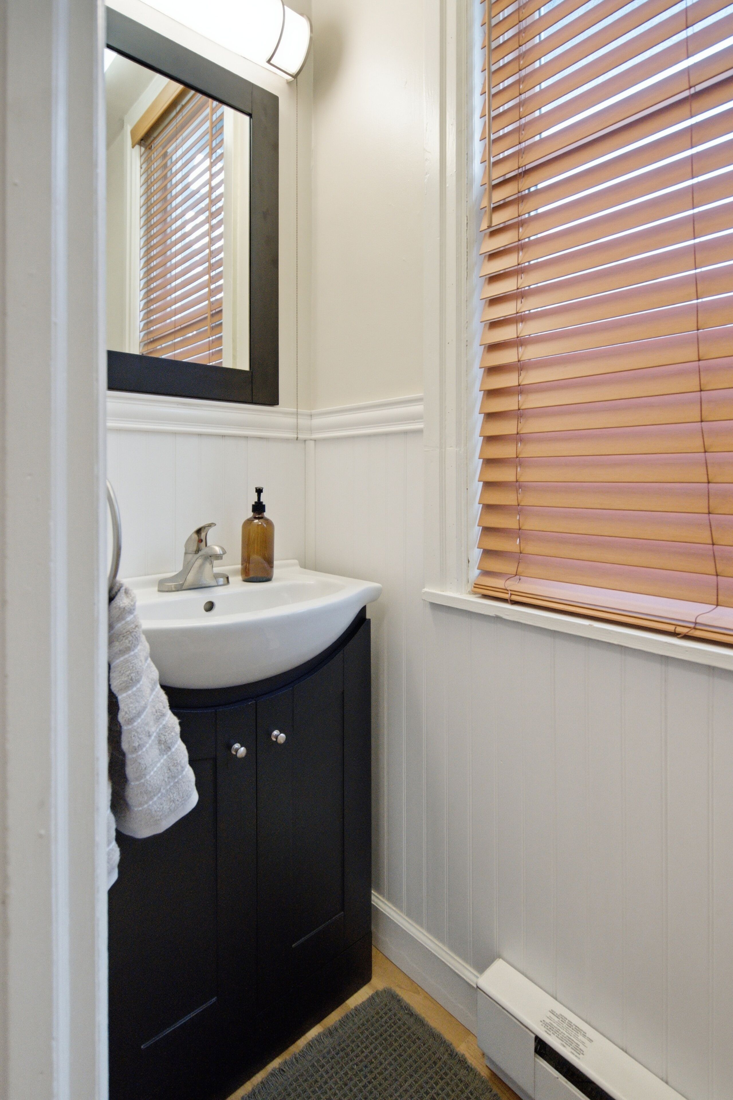 Une salle de bain avec un lavabo blanc, des meubles sombres, un miroir et des stores en bois.