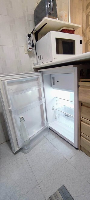 A compact refrigerator with a freezer compartment, a microwave on top, and a power outlet on the wall. - Carlina Canfranc (Canfranc-Estación)
