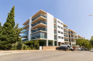 Un immeuble résidentiel à plusieurs étages, doté de balcons, d’un espace de stationnement et entouré de verdure.