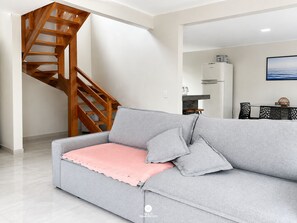 A modern living room with a grey sofa, wooden staircase, and a dining area with a table and chairs. - CasAmar a 50m Praia Pontal do Peró - Cabo Frio/RJ (Cabo Frio)